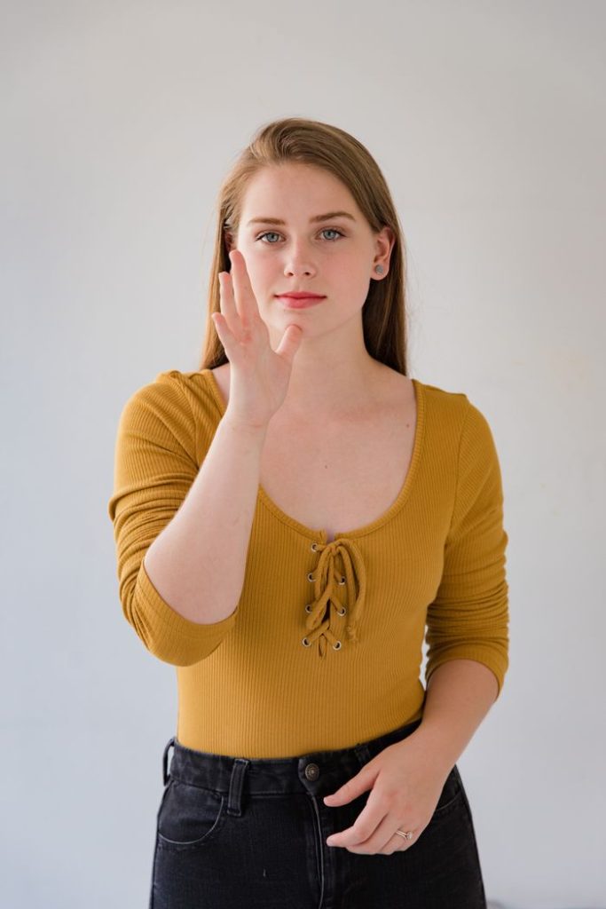 A woman gesturing sign language while facing camera, wearing a yellow top.