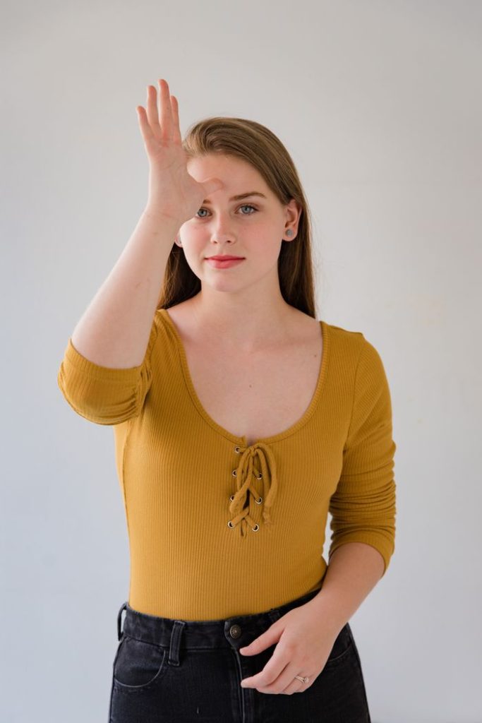 Woman using sign language indoors, wearing mustard blouse, studio setting.